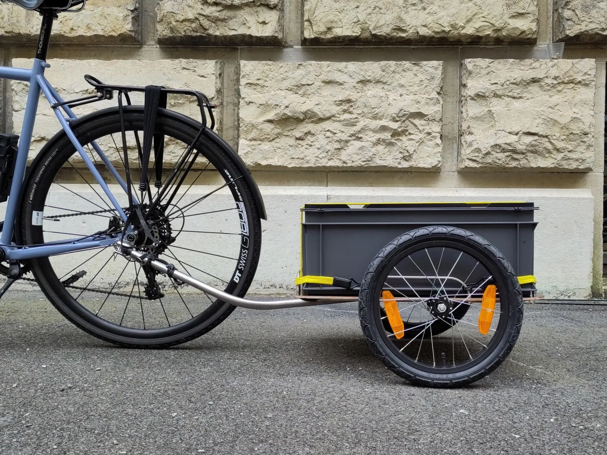 Remorque à caisse grise et châssis en Inox attelée à un vélo, devant un mur de pierre