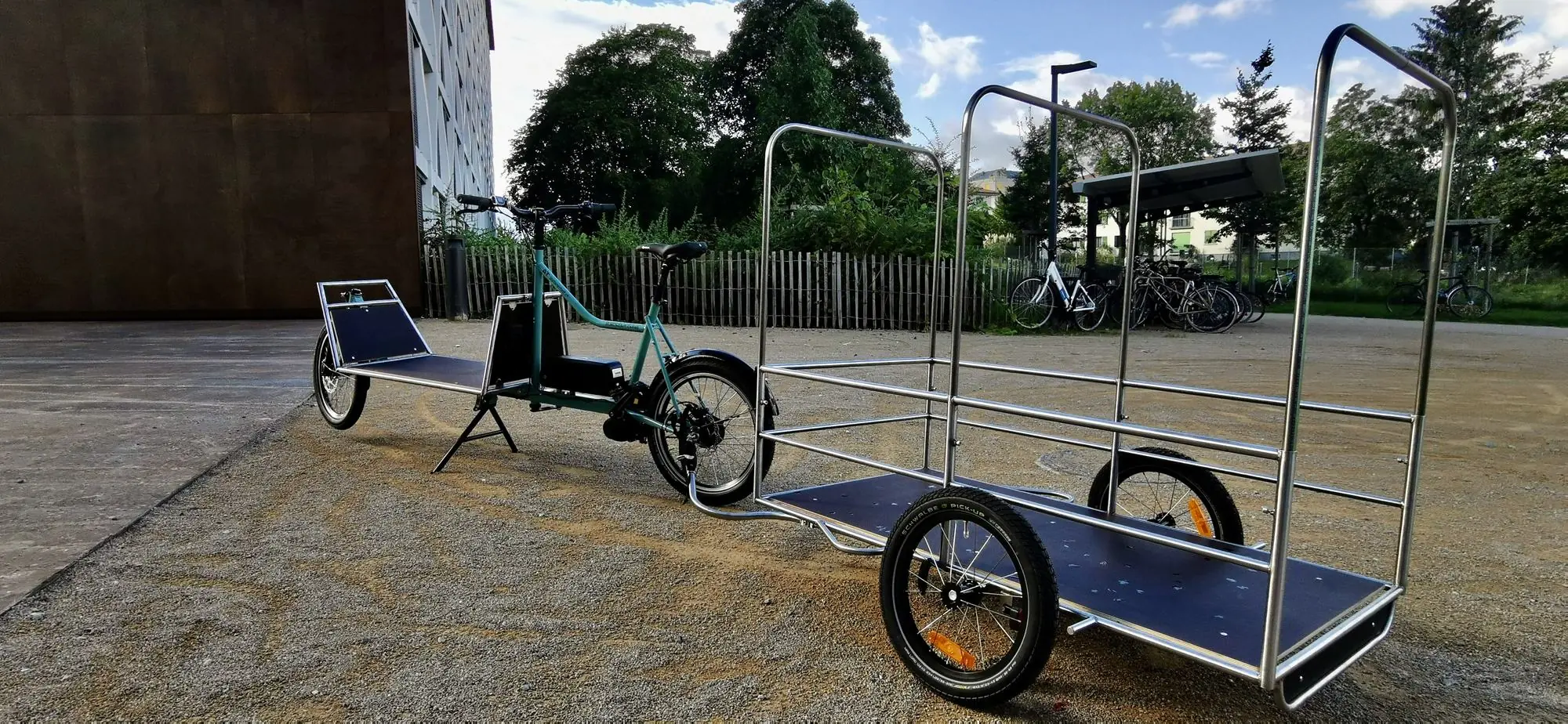 Vélo cargo vert d’eau avec une longue remorque en Inox à haute rambarde, sur la place devant l’atelier