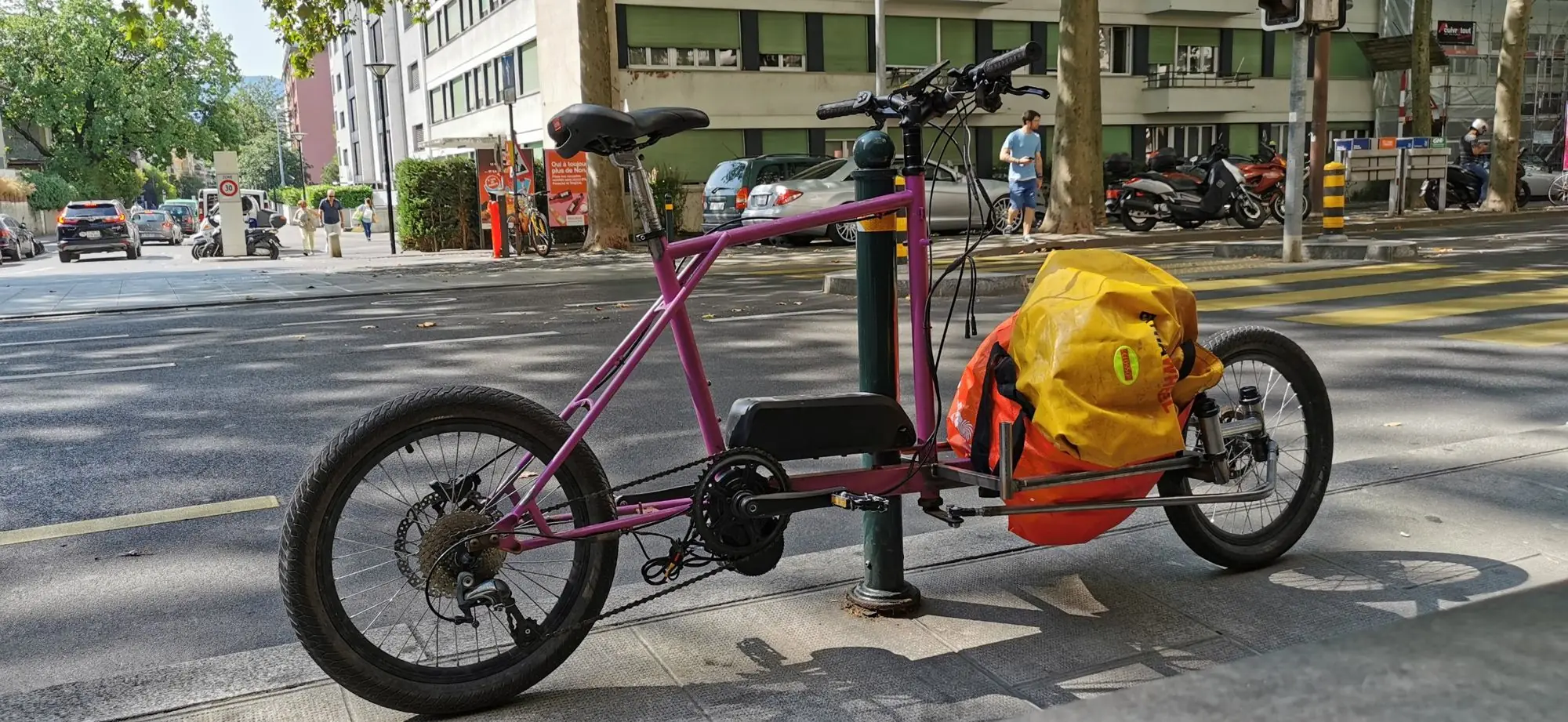 Prototype de vélo cargo longtail violet, sans fourche, garé en ville avec un sac chargé à l’avant